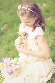 Beautiful smiling little girl playing with cute bunny and sitting on the grass with Easter eggs and flowers in spring holidays. 