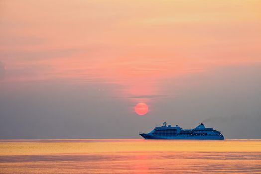 Travel by Cruise ship in the ocean, large luxury passenger boat is sailing on the bright sea, red sun in the colorful sky is yellow, orange, beautiful nature landscape at sunset or sunrise background