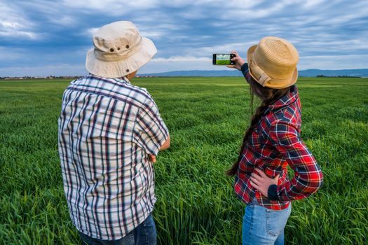 Proud two generations farmers are standing in their barley field in sunset. They are photographing field.