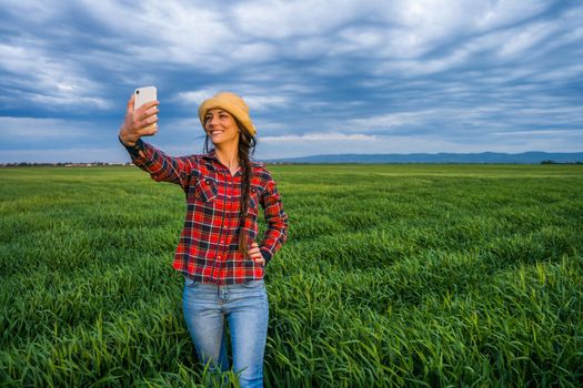 Proud female farmer is standing in her barley field and enjoying sunset. She is taking selfie.