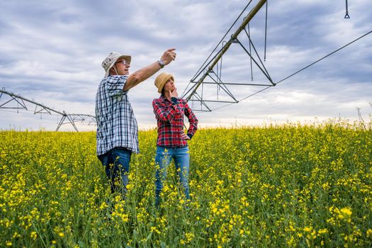 Proud two generations farmers are standing in their rapeseed field. Senior man is education his successor about the farming.