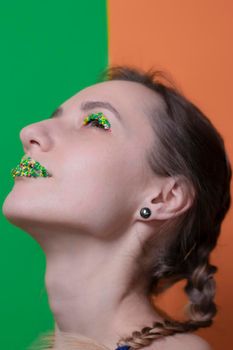 Young woman portrait with a candy makeup - multicoloured pastry topping pearls on her lips and eyelids. On a green and orange background. Easter theme.