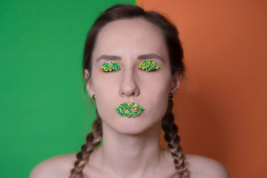 Young woman portrait with a candy makeup - multicoloured pastry topping pearls on her lips and eyelids. On a green and orange background. Easter theme.