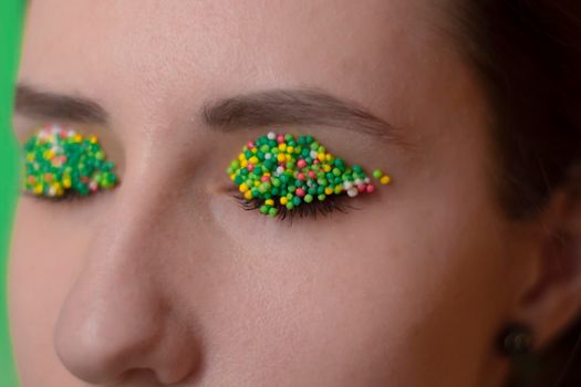 Young woman portrait with a candy makeup - multicoloured pastry topping pearls on her lips and eyelids. On a green and orange background. Easter theme.