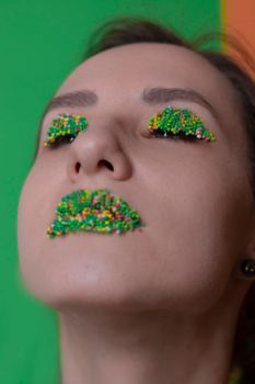 Young woman portrait with a candy makeup - multicoloured pastry topping pearls on her lips and eyelids. On a green and orange background. Easter theme.