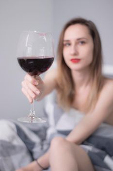 happy beautiful slender half-naked girl shows us a huge glass of red wine, in bed with gray sheets in triangles - vertical photo