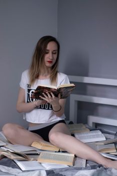 happy beautiful slim girl sitting on the bed with a pile of books, reads. Home schooling. Studying in quarantine. Reading is helpful. Vertical photo