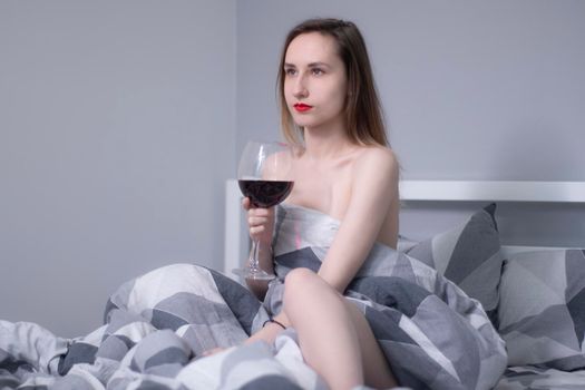 happy beautiful slender half-naked girl holds a huge glass of red wine in bed with gray sheets in triangles - vertical photo