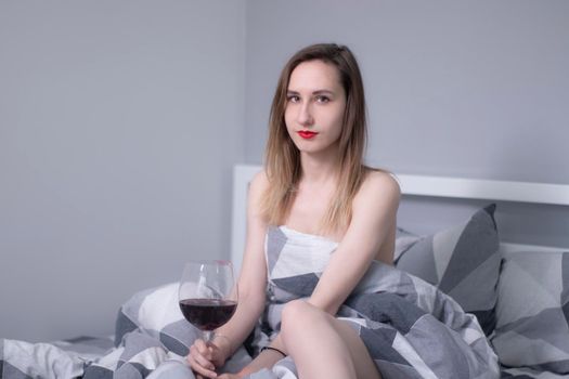 happy beautiful slender half-naked girl holds a huge glass of red wine in bed with gray sheets in triangles - vertical photo