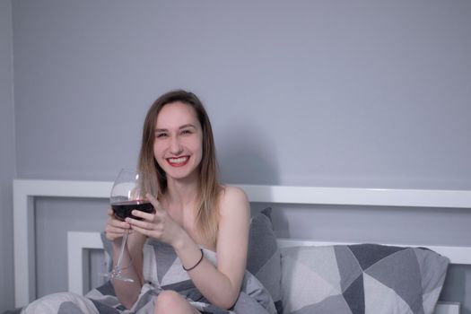 happy beautiful slender half-naked girl holds a huge glass of red wine in bed with gray sheets in triangles - vertical photo