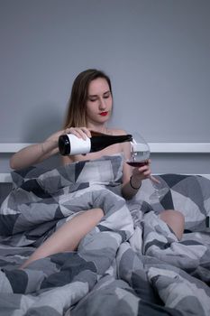 beautiful slender half-naked girl pours red wine from a bottle without a label into a huge glass and in bed- vertical photo