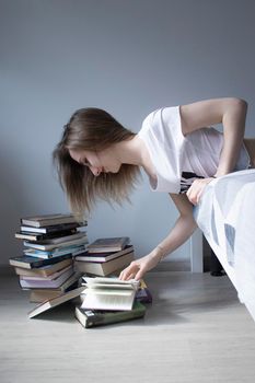 happy beautiful slim girl reading a book lying on the bed with a pile of books nearby. Grey bedroom and sheets. Homeschooling. Studying in quarantine. Reading is helpful. vertical photp