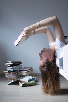 happy beautiful slim girl reading a book lying on the bed with a pile of books nearby. Grey bedroom and sheets. Homeschooling. Studying in quarantine. Reading is helpful. vertical photp