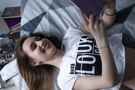 happy beautiful slim girl reading a book lying on the bed with a pile of books nearby. Grey bedroom and sheets. Homeschooling. Studying in quarantine. Reading is helpful. horizontal photp