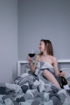 happy slim half-naked girl holds a huge glass of red wine and a bottle without a label in bed - vertical photo