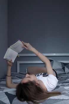 happy beautiful slim girl reading a book lying on the bed with a pile of books nearby. Grey bedroom and sheets. Homeschooling. Studying in quarantine. Reading is helpful. vertical photp