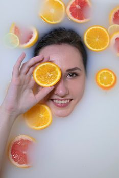 Woman portrait in milk bath with oranges, lemons and grapefruits. Healthy dewy skin. Fashion model girl, spa and skin care concept. Spring colours - yellow, orange, red.