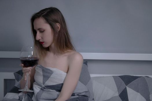 beautiful slender half-naked girl holds a huge glass of red wine in bed with gray sheets in triangles - vertical photo