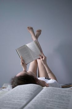 happy beautiful slim girl reading a book lying on the bed full of books, legs up. Grey bedroom and sheets. Homeschooling. Studying in quarantine. Reading is helpful- vertical photo
