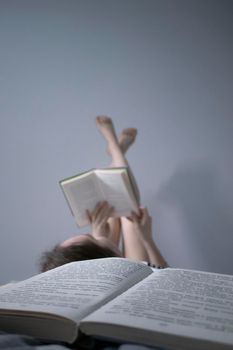 happy beautiful slim girl reading a book lying on the bed full of books, legs up. Grey bedroom and sheets. Homeschooling. Studying in quarantine. Reading is helpful- vertical photo