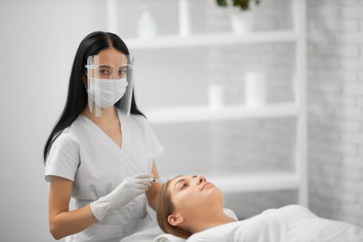 Side view of young beautician in white uniform and protective mask holding syringe and doing beauty injection for skin or hair. Concept of procedure for hair restoration and improvements growth.