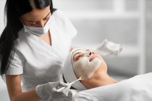 Side view of beautiful brunette beautician applying special white cream for face young cute patient. Concept of process procedure for improving face skin with cosmetics.