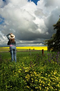 Country woman in jeans at the farm gate watching the rains come in over field crops of canola