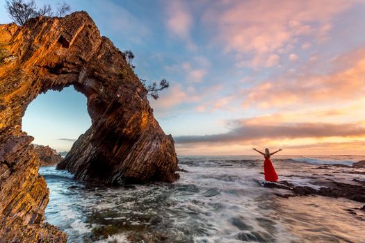 Woman in long flowing red dress standing in ocean waves by an incredible sea arch.  Her dress in motion with the waves