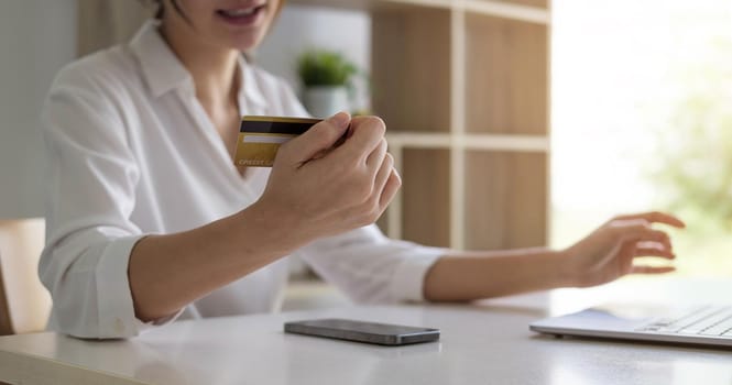 Attractive Asian woman sitting relaxed on sofa, holding credit card and smartphone probably buying something online..