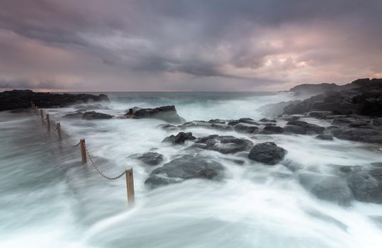 Stormy weather and large swell waves wash over rocks into rock pool, the heavy chains tossed by the force of water as well as some of the looser rocks which make deep  creaking noises.  Note a 4 second exposure showing deliberate motion
