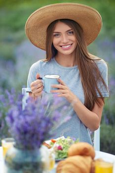 Portrait of young stylish woman holding cup of coffee, posing at table with dinner outdoors. Gorgeous girl wearing straw hat and dress sitting and looking at camera, lavender field on background.