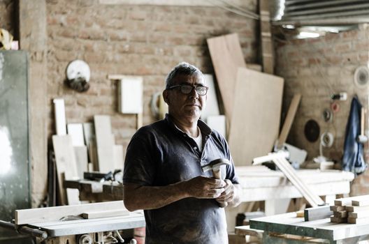 Portrait of senior carpenter. Standing in his workshop and looking at camera.