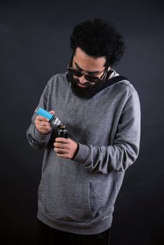 young vaper man with beard vaping mechanical mod. Guy smokes an electronic cigarette by blowing a smoke vapor. Holds in hand Refills rda juice from a bottle