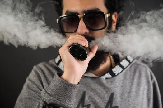 Portrait of a young guy with big beard and sunglasses and vaping an electronic cigarette on a black background.