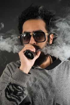 Portrait of a young guy with big beard and sunglasses and vaping an electronic cigarette on a black background.