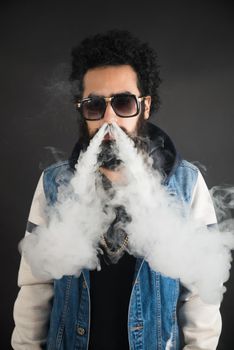 Young man vaping, studio shot. Bearded guy with sunglasses blowing a cloud of smoke on black background. Concept of smoking and steam without nicotine, copy space