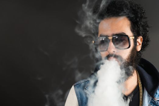 Young man vaping, studio shot. Bearded guy with sunglasses blowing a cloud of smoke on black background. Concept of smoking and steam without nicotine, copy space