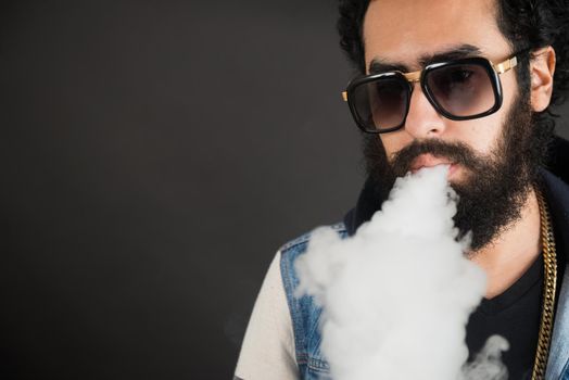 Young man vaping, studio shot. Bearded guy with sunglasses blowing a cloud of smoke on black background. Concept of smoking and steam without nicotine, copy space