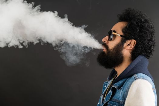 Young man vaping, studio shot. Bearded guy with sunglasses blowing a cloud of smoke on black background. Concept of smoking and steam without nicotine, copy space