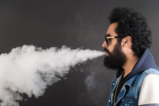 Young man vaping, studio shot. Bearded guy with sunglasses blowing a cloud of smoke on black background. Concept of smoking and steam without nicotine, copy space