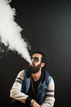 Young man with beard vaping an electronic cigarette upwards. Vaper hipster smoke vaporizer. Black background