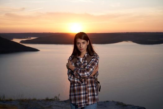 Front view of attractive woman in checkered shirt standing on high hill and looking at camera. Amazing sunset over Dniester river on background.