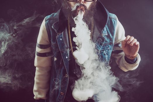 Young man with beard throwing a cloud of steam. Black background. Vaping an electronic cigarette with a lot of smoke