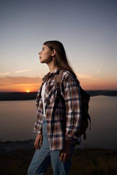 Charming dark-haired woman in checkered shirt walking with backpack at Bakota bay during amazing sunset. Concept of vacation and adventure.