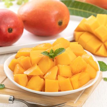 Diced fresh mango fruit on a white plate with leaves over wooden table background.