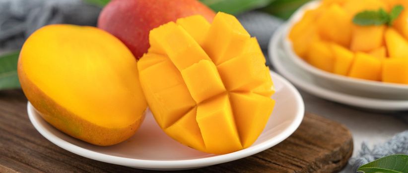 Diced fresh mango fruit on a white plate over wooden cutting board with leaves on gray table background.