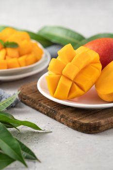 Diced fresh mango fruit on a white plate over wooden cutting board with leaves on gray table background.