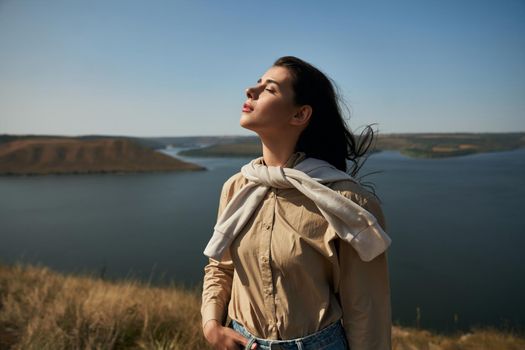 Pleasant woman with dark hair standing high with closed eyes with amazing view of Dniester river on background. Bakota area for hiking.