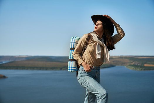 Beautiful young woman in cowboy hat standing on peak of mountain with closed eyes. Scenic view of Dniester river on background. Natural landscape.