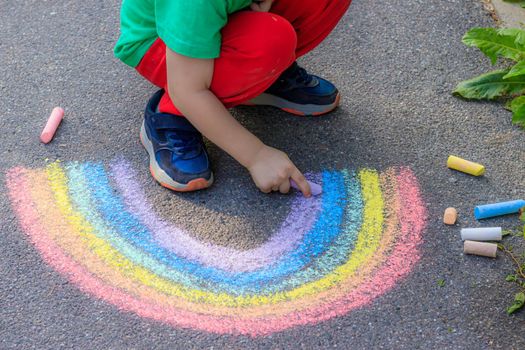 A boy draws a rainbow on the asphalt with crayons . A child draws on the asphalt . Children's drawing. Walking in the city with a child. Summer in the city. Crayons for drawing. Article about childhood.